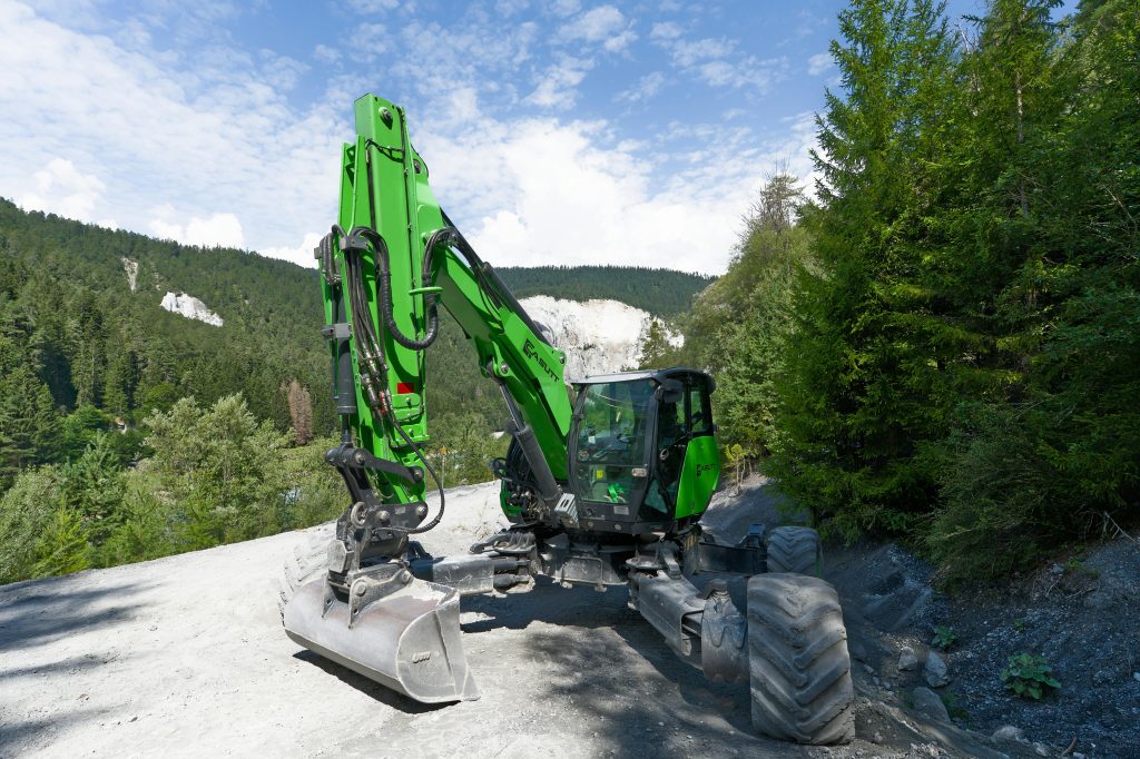 A green excavator operating in a scenic Swiss alpine forest with clear skies.